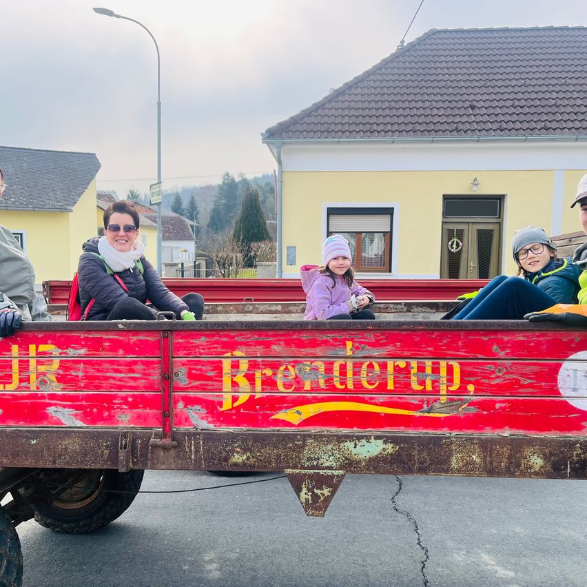 Vier Personen, darunter ein junges Mädchen, sitzen auf der Rückseite eines roten Trucks mit dem Wort Brederup darauf. Sie scheinen sich in einem Dorf mit einem Haus im Hintergrund zu befinden.
