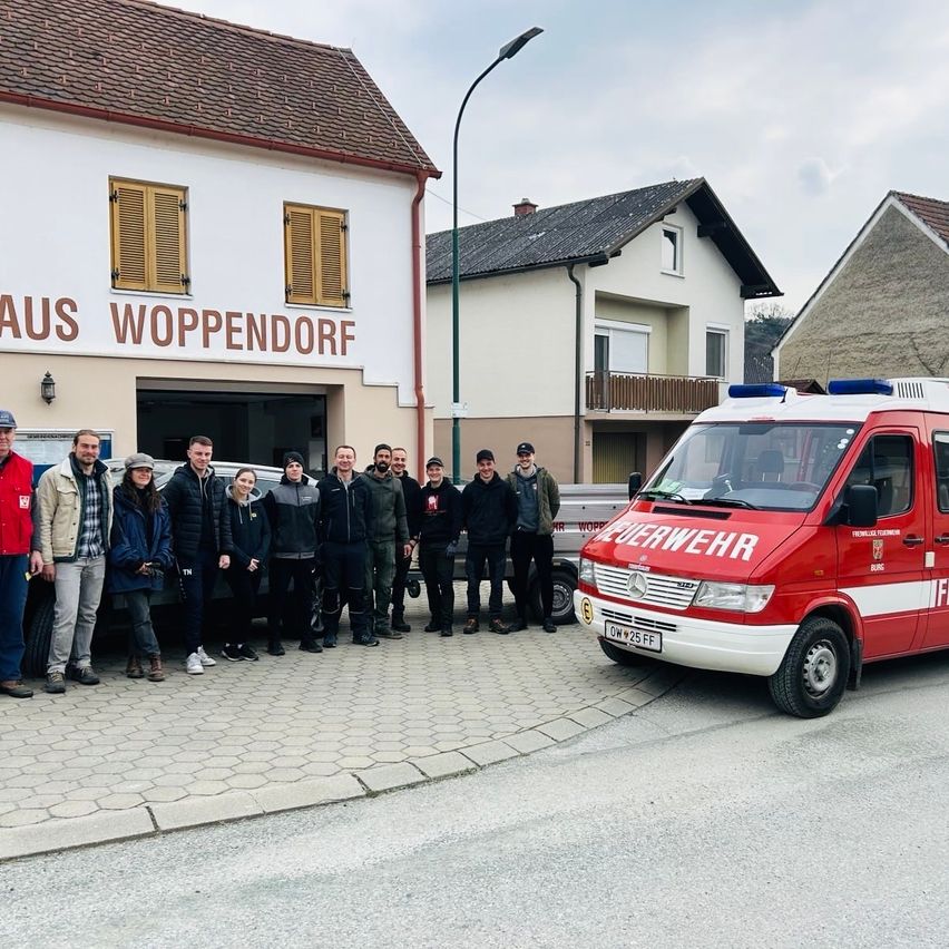 Eine Gruppe von Menschen steht vor einer Feuerwehrwache in Woppendorf, mit einem geparkten Krankenwagen in der Nähe.