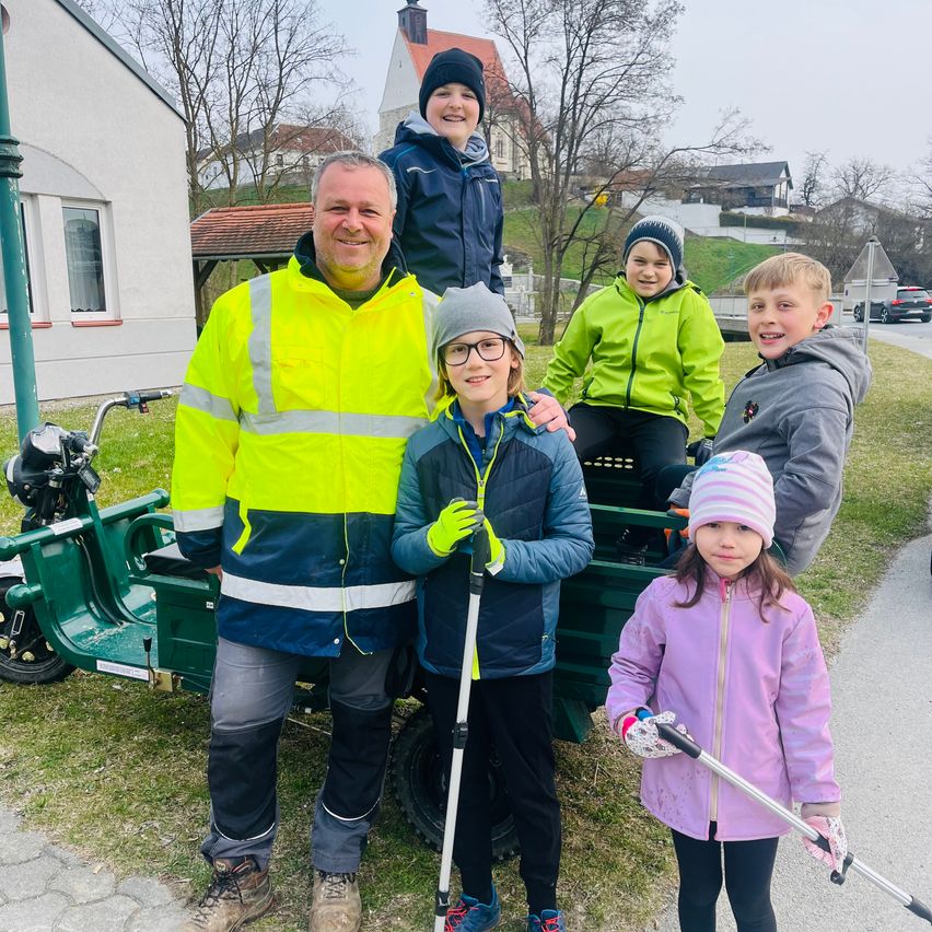 Eine Familie mit fünf Mitgliedern, einschließlich eines Mannes und vier Kindern, steht draußen neben einem grünen Fahrzeug und hält Besen in der Hand und lächelt. Sie sind in warme Jacken gekleidet und scheinen bereit für eine Aktivität zu sein.