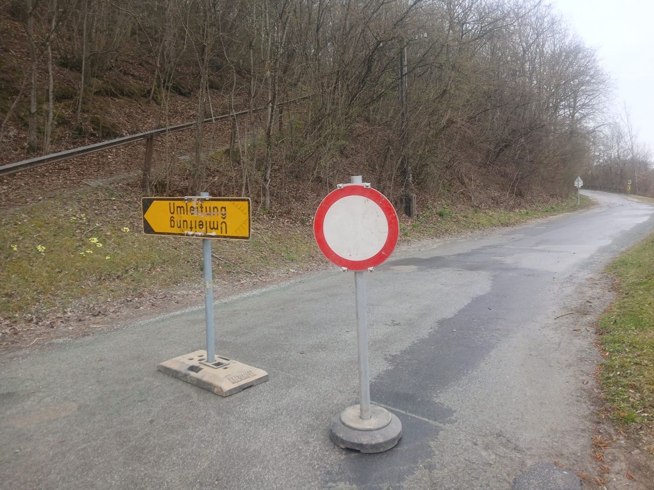 Eine Straße mit zwei Schildern; eines ist gelb mit schwarzem deutschem Text, das andere ein rundes rot-weißes Schild. Die Straße ist nass.