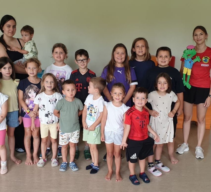 A group of children and adults posing for a photo, with a woman holding a baby and a frog toy in the background.