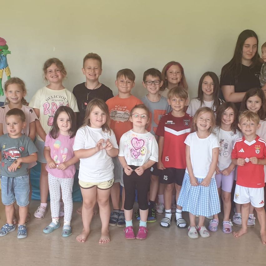 A group of children and two adults are posing for a photo in a room with a yellow wall. The children are standing in a row, smiling, and wearing various outfits. The adults are standing behind them, one holding a baby.