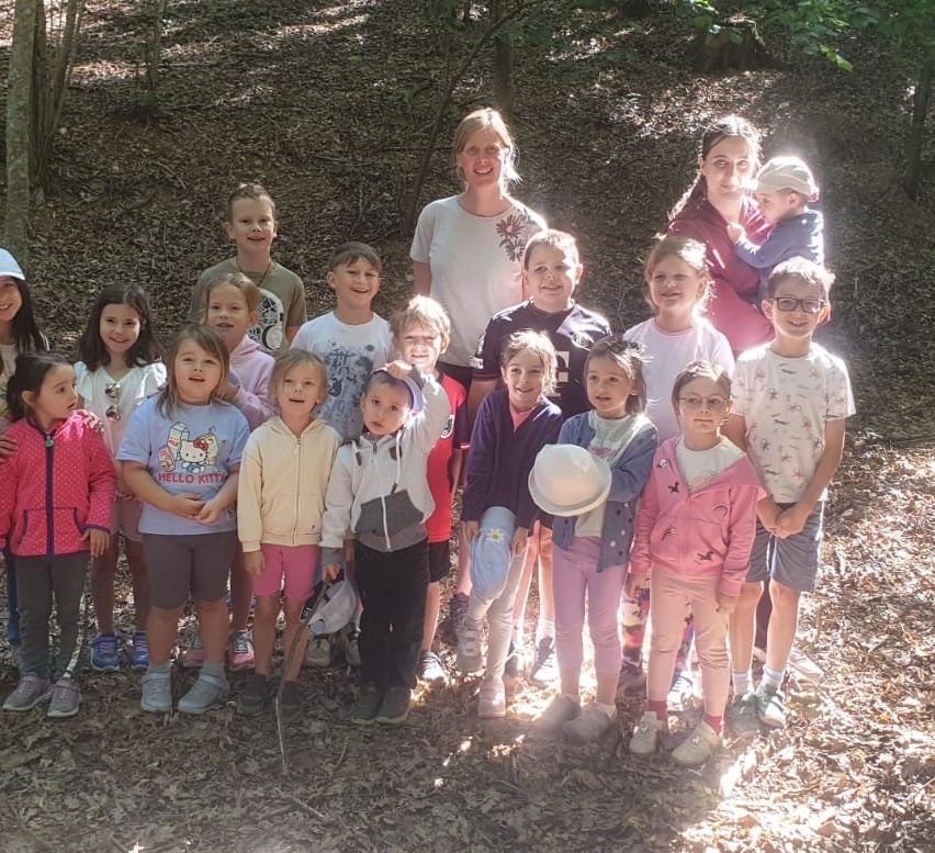 A group of children and two adults stand in a forest, smiling for a photo. The children wear various jackets and sneakers, while the adults wear hats.