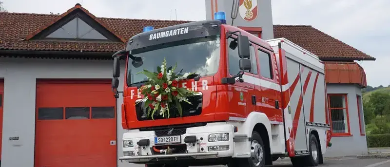 Ein roter Feuerwehrwagen mit dem Wort Baumgartner steht vor einer Feuerwache. Er hat Blumen auf der Vorderseite.