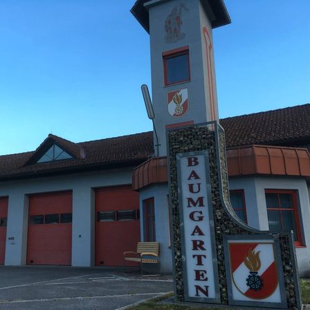Die Vorderseite einer Feuerwache hat ein Schild mit der Aufschrift 'Baumgarten'. Das Gebäude hat ein rotes Dach und rote Türen. Ein Stuhl ist davor aufgestellt.