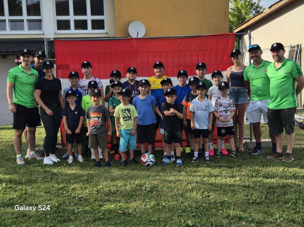 A group of children in matching caps pose for a photo in front of a red banner, with adults behind them and a soccer ball on the ground.