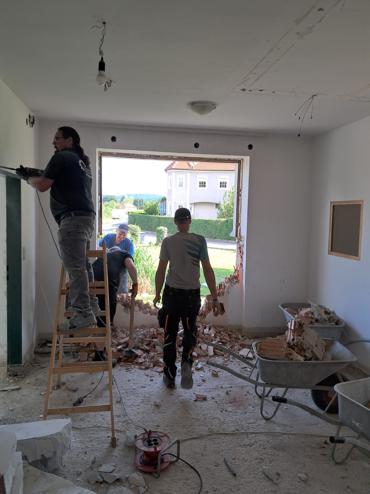 Bauarbeiter mit Bohrhammer auf Leiter in einem Raum mit Bauschutt, Schaufel und Schubkarren