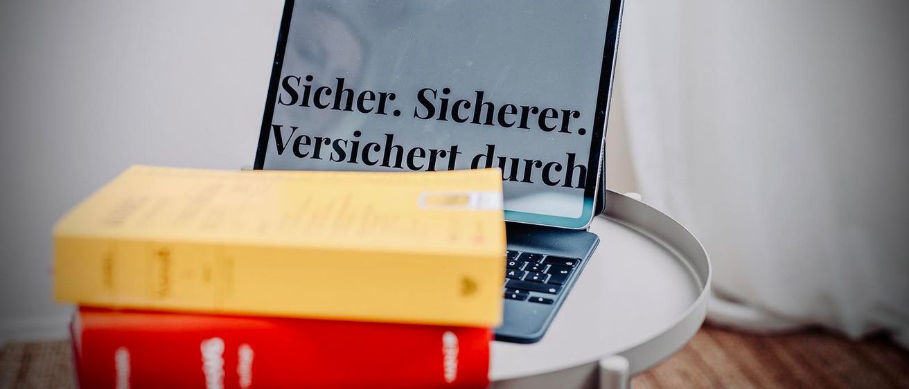 Ein Laptop mit einem Bildschirm, auf dem 'Sicher. Sicherer. Versichert durch' steht, steht auf einem runden Tisch. Zwei Bücher, eines gelb und eines rot, liegen auf dem Laptop.
