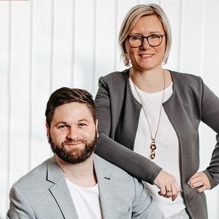 Zwei Personen, eine Frau mit Brille und ein Mann mit Bart, sitzen zusammen in einem Studio. Die Frau steht hinter dem Mann, mit ihrem Arm auf seiner Schulter. Beide lächeln für die Kamera.