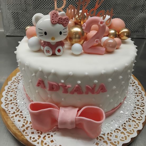 Ein Geburtstagskuchen dekoriert mit Hello Kitty, goldenen und rosa Luftballons und der Nummer 2 auf der Oberseite. Der Kuchen hat ein weißes und rosa gepunktetes Design. Der Kuchen steht auf einem Holzteller mit einer rosa Schleife.