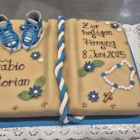 Ein Kuchen in Form eines offenen Buches mit Babyschuhen, Blumen und einer Kette. Die Namen 'Fabio' und 'Lorian' sind in Braun geschrieben. Das Datum '8.Juni2015' steht auf dem Kuchen. Der Kuchen liegt auf einem Spitzentuch.