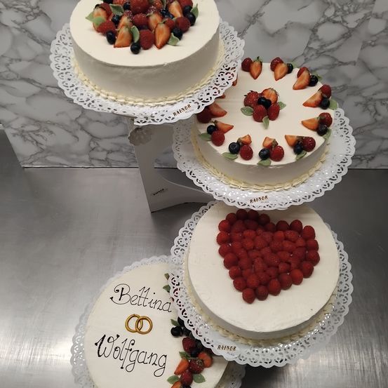 Drei Kuchen auf einem dreistufigen Kuchenständer. Der oberste Kuchen hat weiße Glasur und eine Mischung aus Erdbeeren und Blaubeeren. Der mittlere Kuchen hat die Form eines Herzens aus Himbeeren. Der unterste Kuchen ist mit einer Hochzeitsringe und den Namen 'Bettina' und 'Wolfgang' verziert.