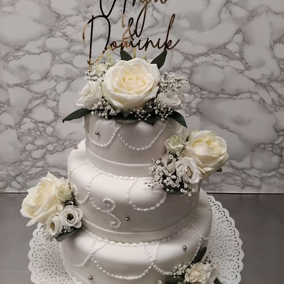 Ein dreistöckiger weißer Hochzeitskuchen mit weißen Rosen und Babys Atmung Blumen. Die oberste Etage hat ein goldenes Namensschild mit 'Mygo & Dominik'. Der Kuchen steht auf einem Doily auf einer Marmorfläche.