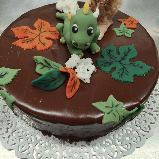 Ein Schokoladenkuchen verziert mit einem grünen Drachen, Blättern und kleinen weißen Toppings, auf einem silbernen Doily.