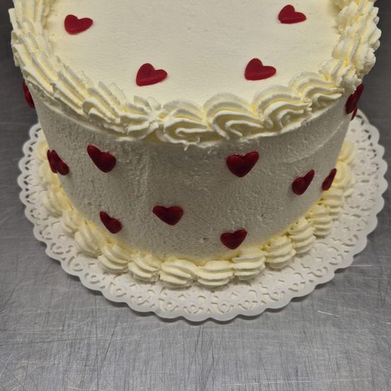 Ein weißer Kuchen mit cremiger Glasur und roten Herzdekorationen auf einer Spitze auf einer silbernen Oberfläche.