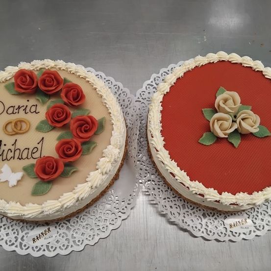 Zwei Kuchen auf Spitzeplatten. Der linke Kuchen hat rote Rosen und ein Herz, mit den Namen Daria und Michael in Weiß geschrieben. Der rechte Kuchen hat drei weiße Rosen auf einer roten Basis. Beide haben weiße Glasur.