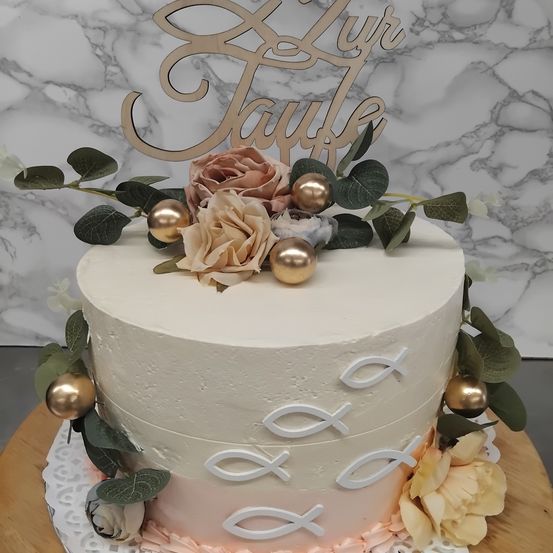 Ein dreischichtiger Kuchen mit weißer Glasur und dekorativen Fischdesigns, gekrönt von einem personalisierten Holzschild und Blumen. Er steht auf einer Holzunterlage vor einem Marmorhintergrund.