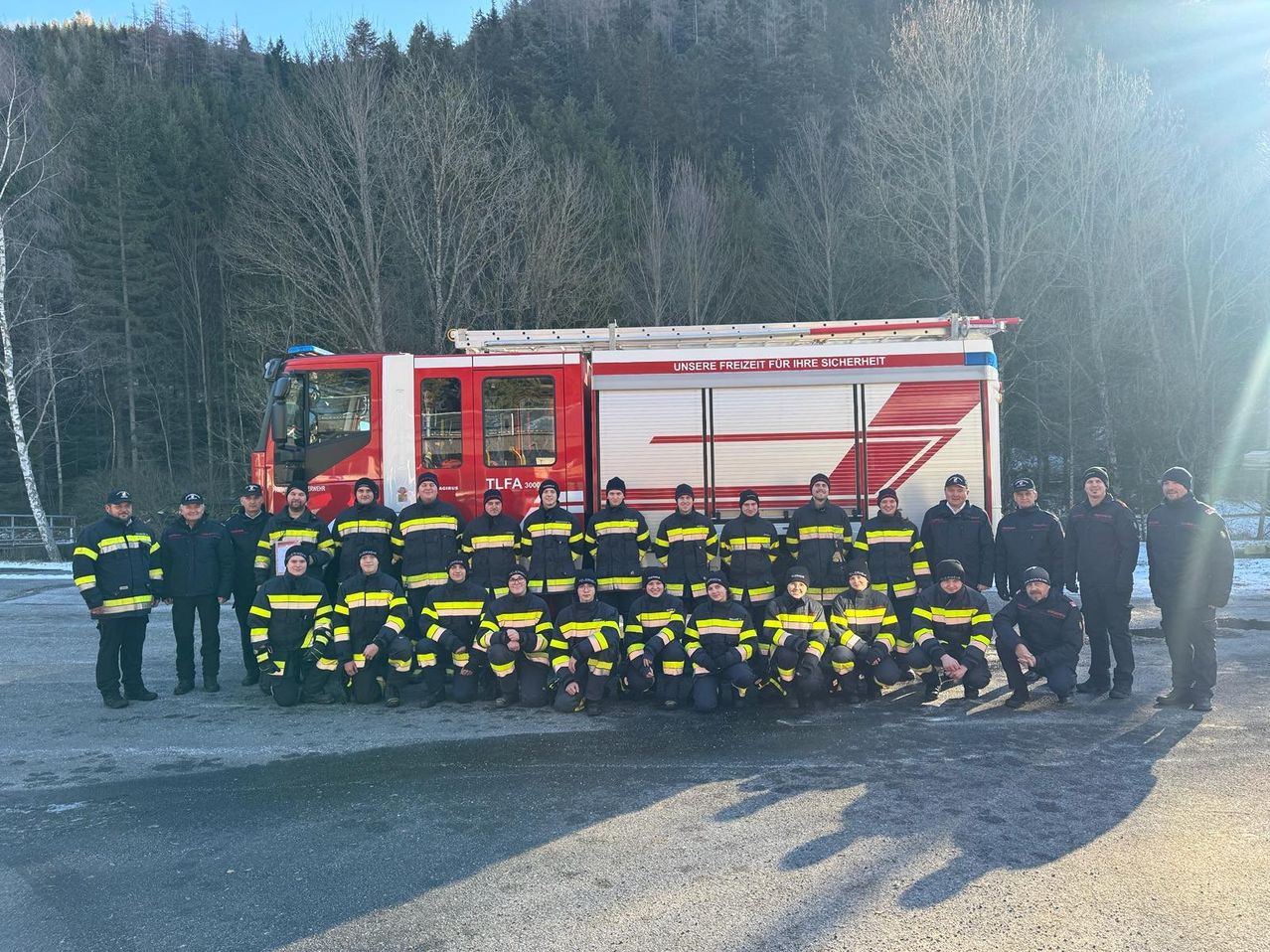 Eine Gruppe Feuerwehrleute in passenden Uniformen posiert für ein Foto vor einem Feuerwehrwagen. Sie sind von einem Waldhintergrund umgeben.