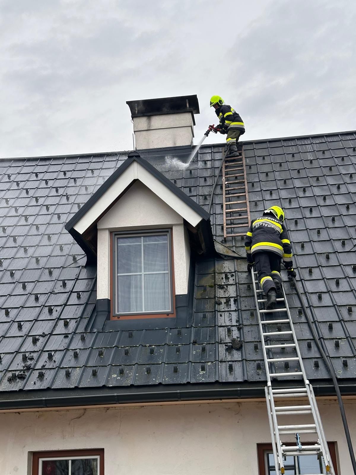 Zwei Feuerwehrleute sind auf einem Dach und reinigen es. Einer benutzt einen Schlauch, während der andere eine Leiter erklimmt. Das Dach hat einen Schornstein und ein Fenster.