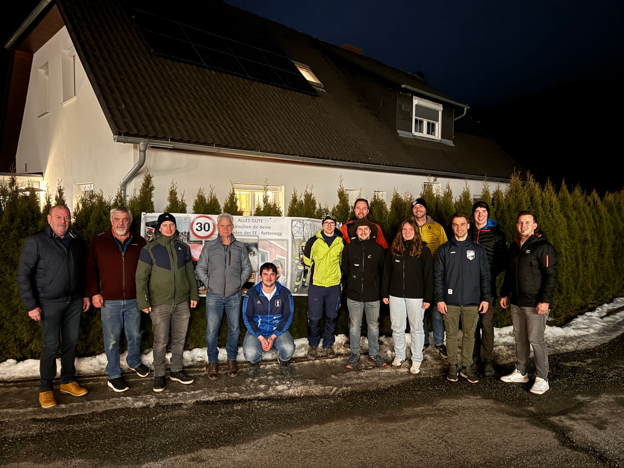 A group of people stands in front of a house with solar panels at night. They are dressed in winter clothing and some are kneeling. A signboard and a 30 km/h speed limit sign are behind them.