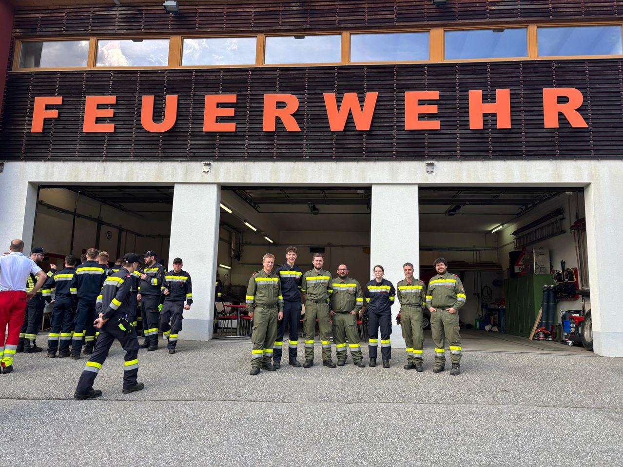 Eine Gruppe Feuerwehrleute steht vor einem Gebäude mit der Aufschrift Feuerwehr. Sie tragen Uniformen und einige schauen zur Seite.