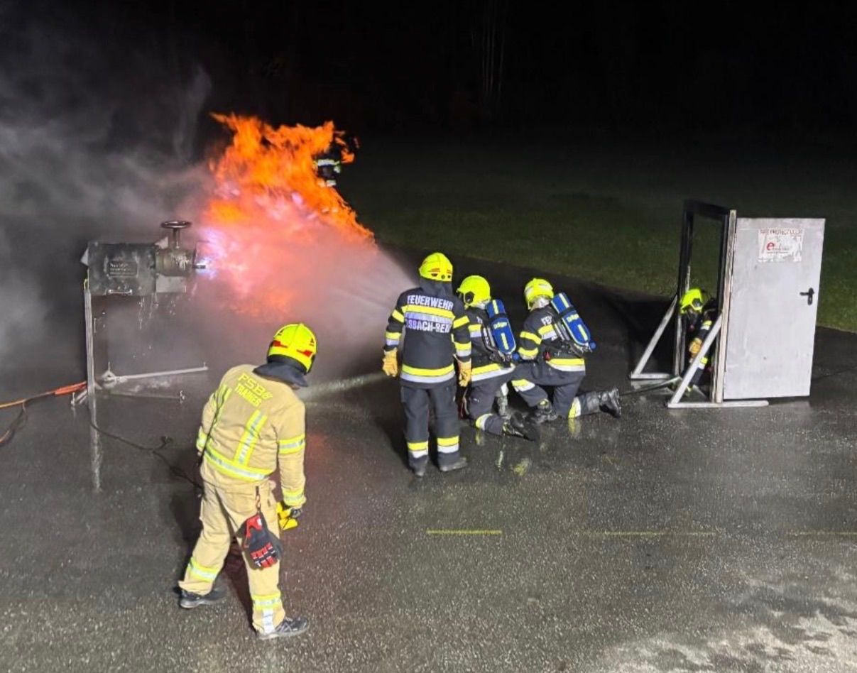 Firefighters are using a hose to extinguish a fire on a pump. One firefighter kneels while another stands ready.