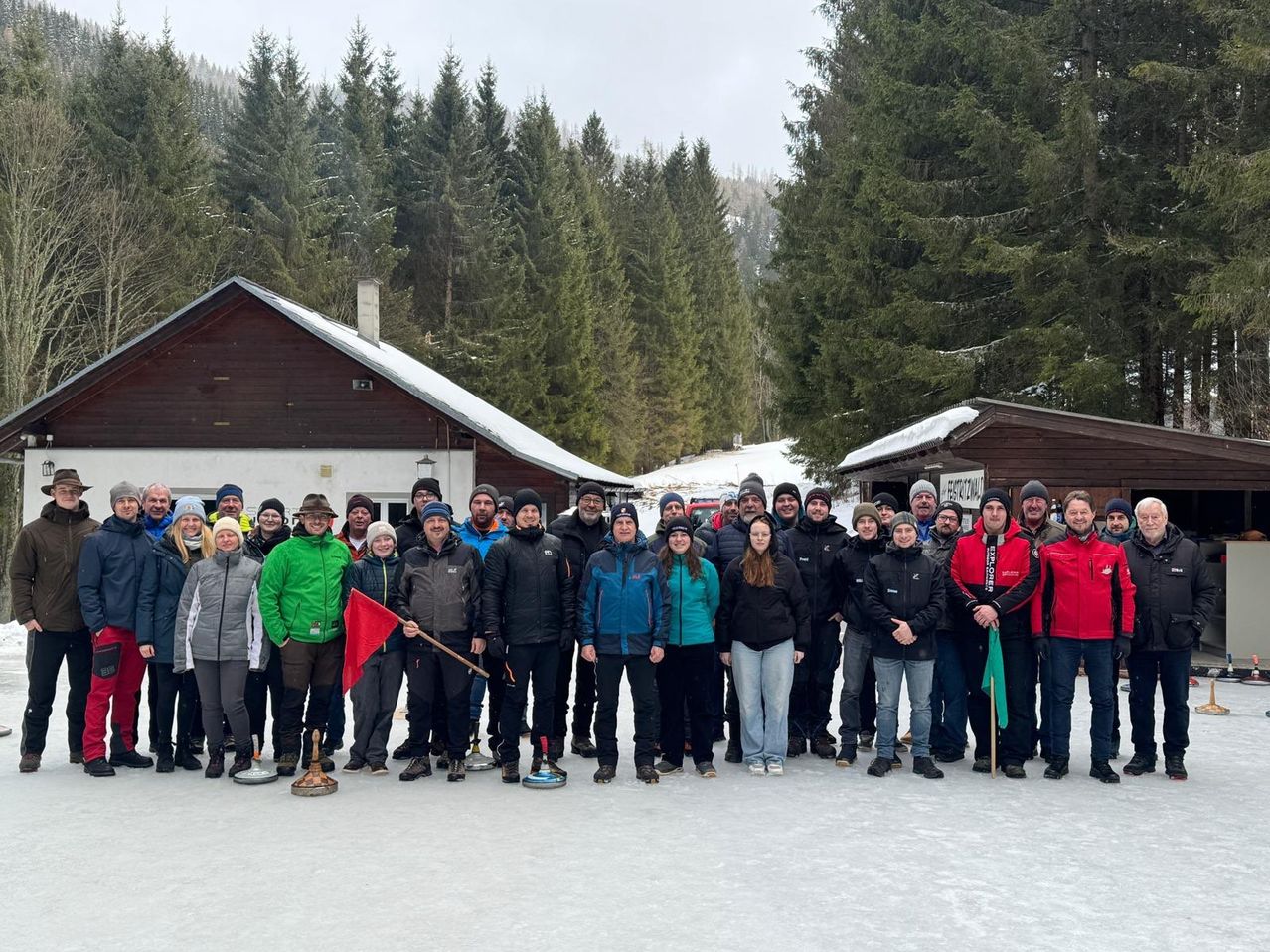 Eine Gruppe von Menschen posiert für ein Foto auf einer Eisbahn, umgeben von verschneiten Kiefern und kleinen Gebäuden. Sie tragen Winterkleidung und halten Stöcke.