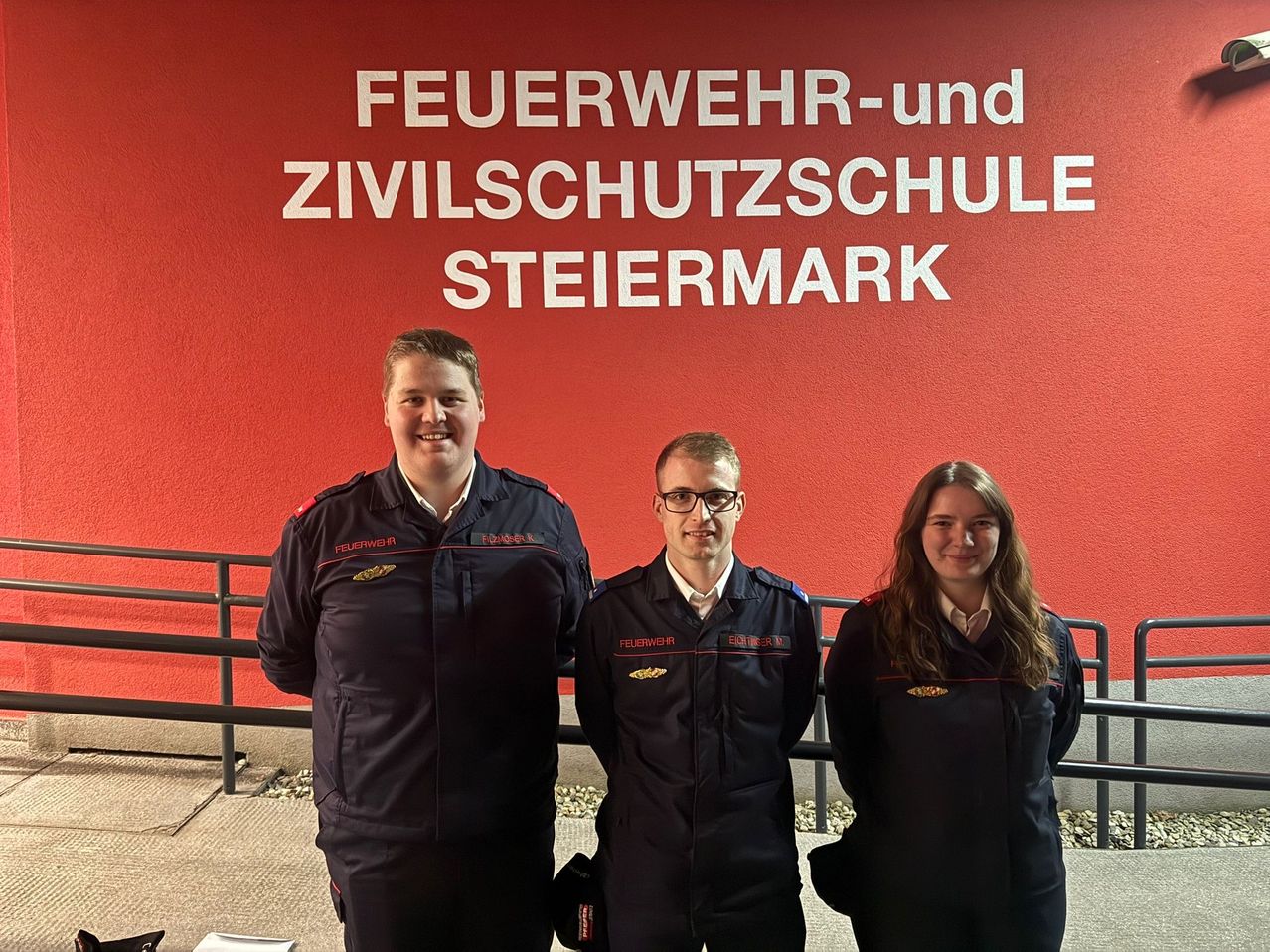 Drei Personen in gleichen Uniformen stehen vor einer roten Wand mit weißem Text. Sie scheinen für ein Foto zu lächeln.