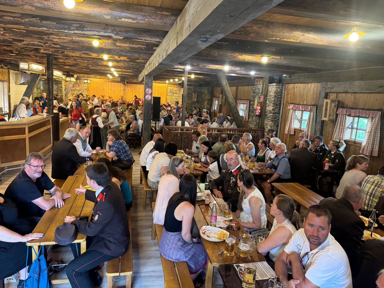 Eine große Gruppe von Menschen sitzt an Holztischen in einem rustikalen Restaurant, einige in Uniformen, mit Tellern Essen, Getränken und einer warmen Atmosphäre.