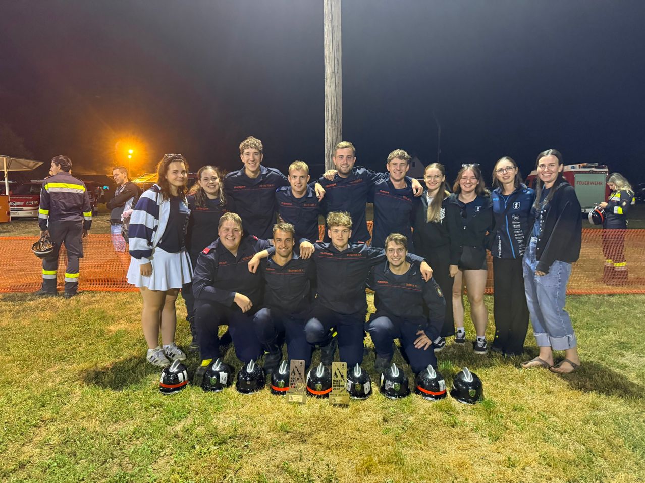 A group of people in matching uniforms pose for a photo at night, with helmets and a trophy in front of them.