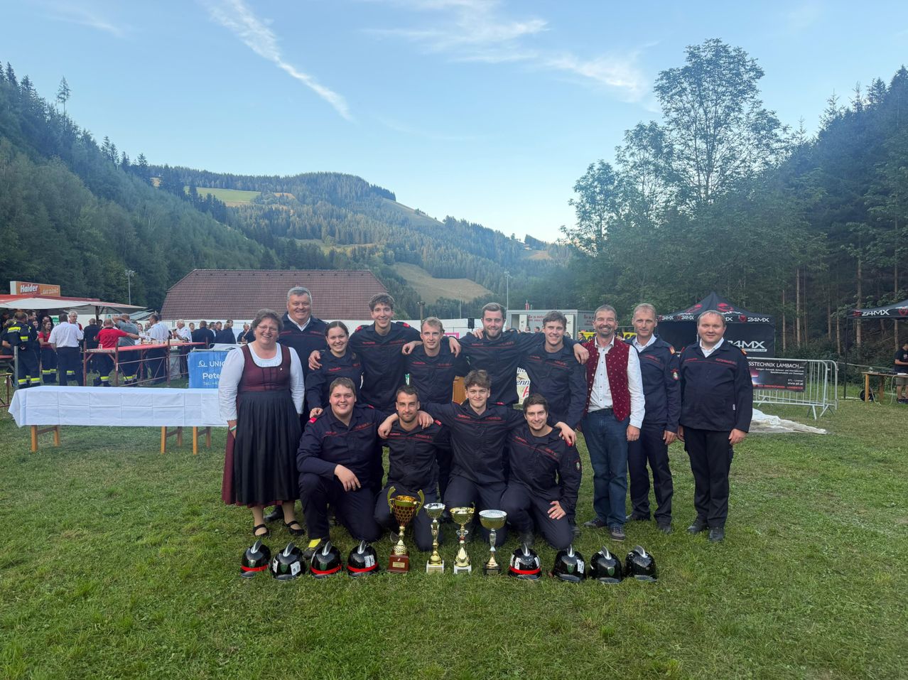 A group of people dressed in matching uniforms pose for a photo on a grassy field with mountains in the background. They are holding trophies and helmets.