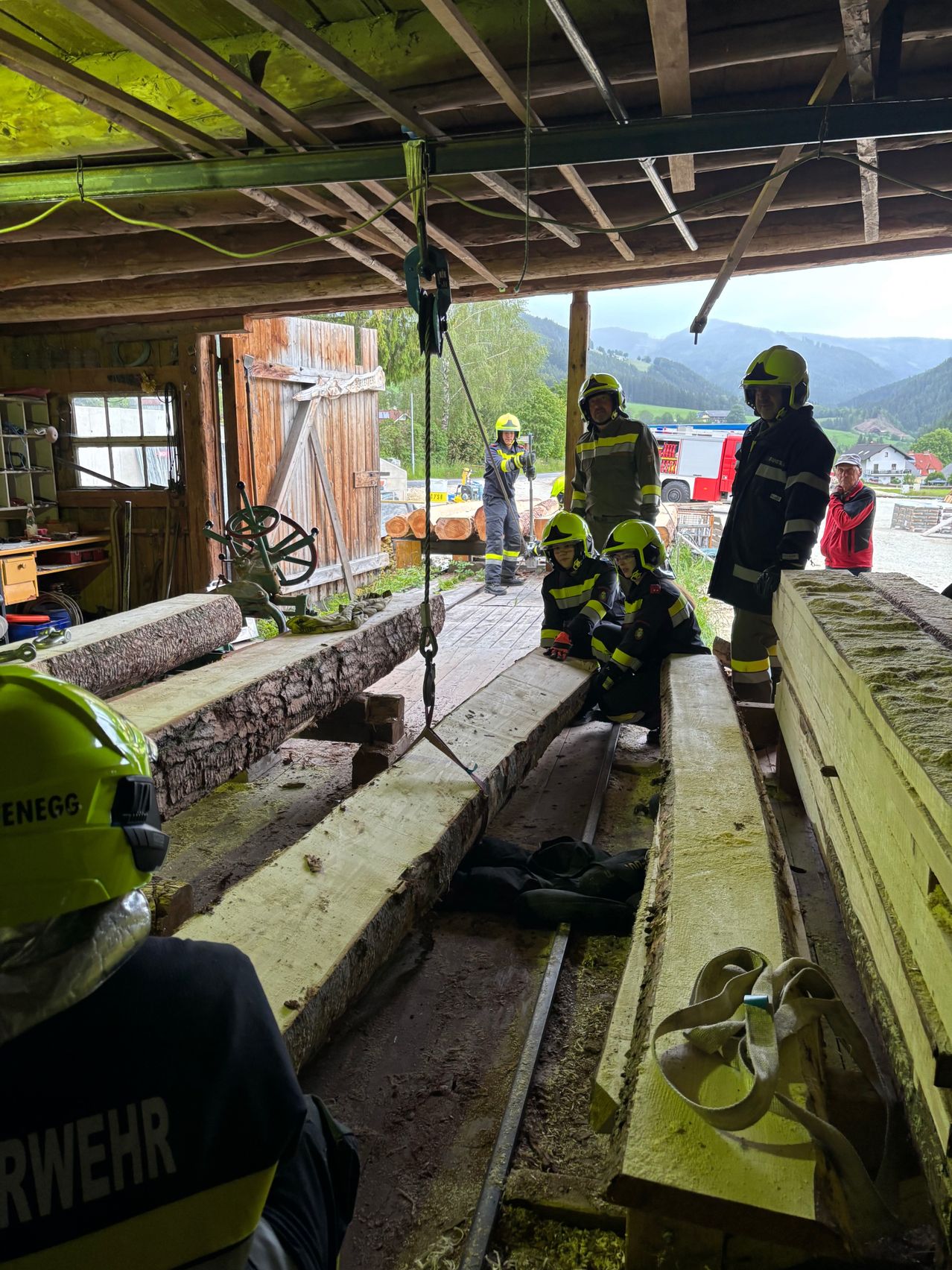 Eine Gruppe von Feuerwehrleuten in Schutzausrüstung arbeitet mit großen Holzbalken in einer Scheune mit einem bergigen Hintergrund.