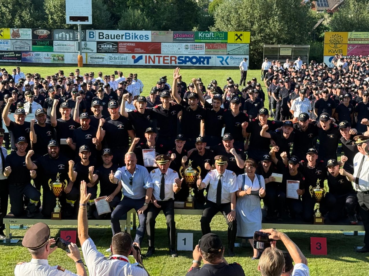 Eine große Gruppe von Menschen in Uniform, wahrscheinlich Polizeibeamte, feiert auf einem Rasenfeld. Sie posieren mit ihren Trophäen, und einige heben die Hände. In der Mitte sitzen einige auf einer Bank, während andere Fotos machen.