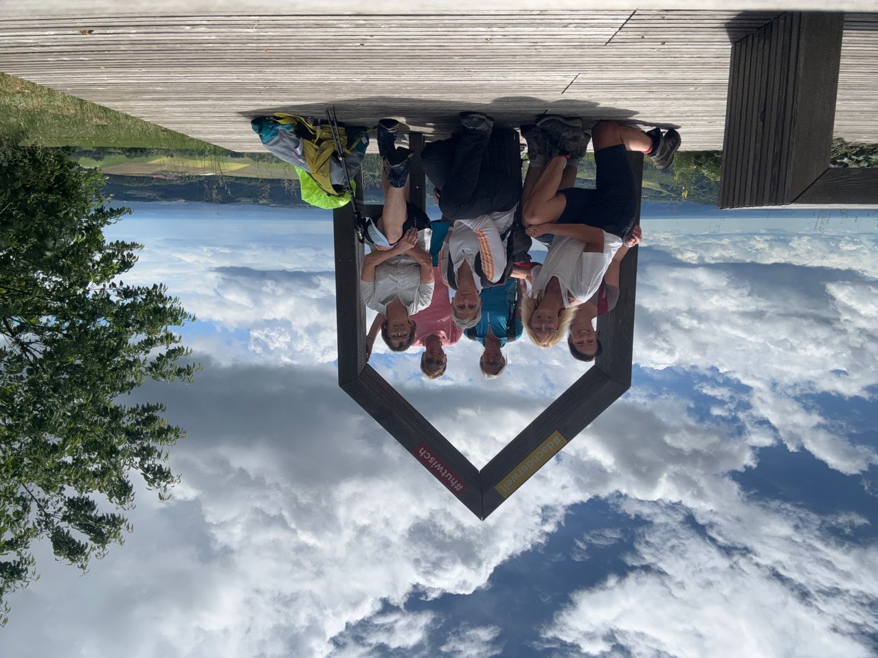Sieben Menschen in Wanderausrüstung posieren in Herzform auf einer Holzbrücke mit Wolken darunter.
