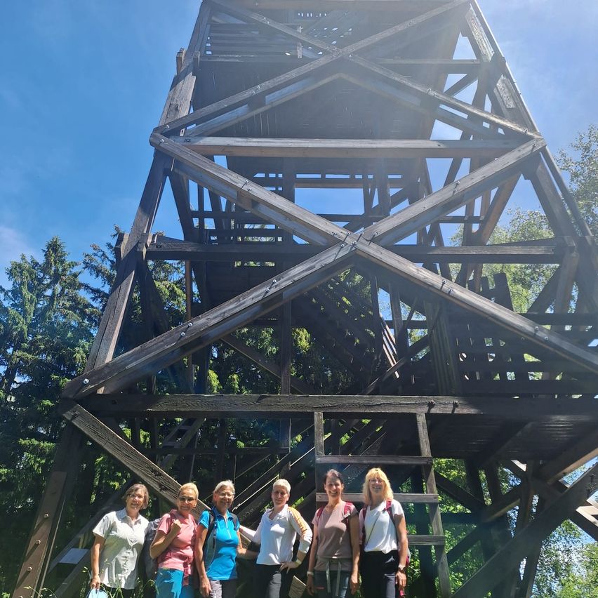 Fünf Frauen in Wanderausrüstung stehen gemeinsam vor einem alten Holzturm unter einem strahlend blauen Himmel.