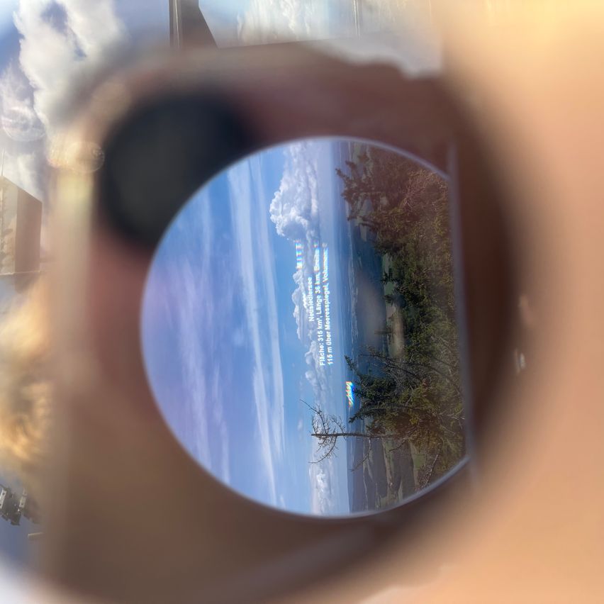 Eine szenische Ansicht durch eine Sonnenbrille mit Strand, Bäumen und einem Berg mit einer Wolke. Der Himmel ist blau mit einigen Wolken.