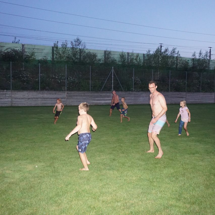 Eine Gruppe von Menschen, darunter Erwachsene und Kinder, spielt auf einer Rasenfläche unter einem Dämmerungshimmel, wobei Stromleitungen im Hintergrund sichtbar sind.