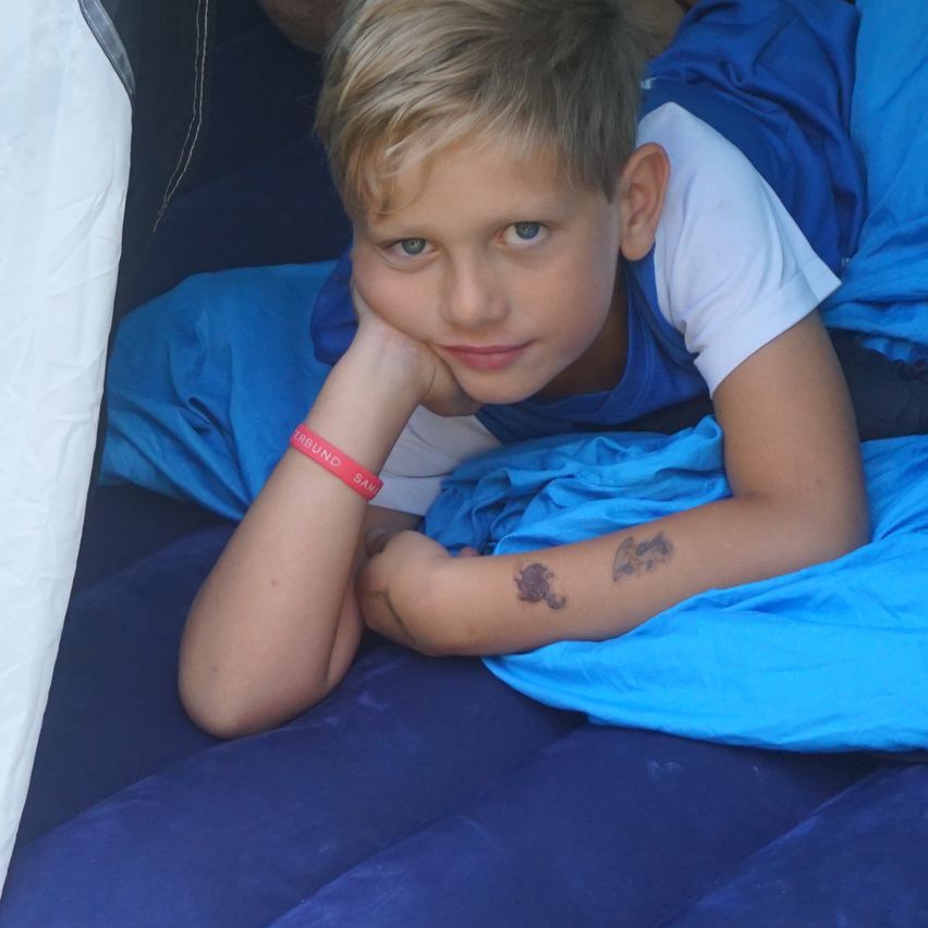 Ein junger Junge mit blonden Haaren liegt auf einem blauen Schlafsack, trägt ein blaues und weißes T-Shirt und hat ein rosa Armband und zwei kleine Tätowierungen am linken Arm.