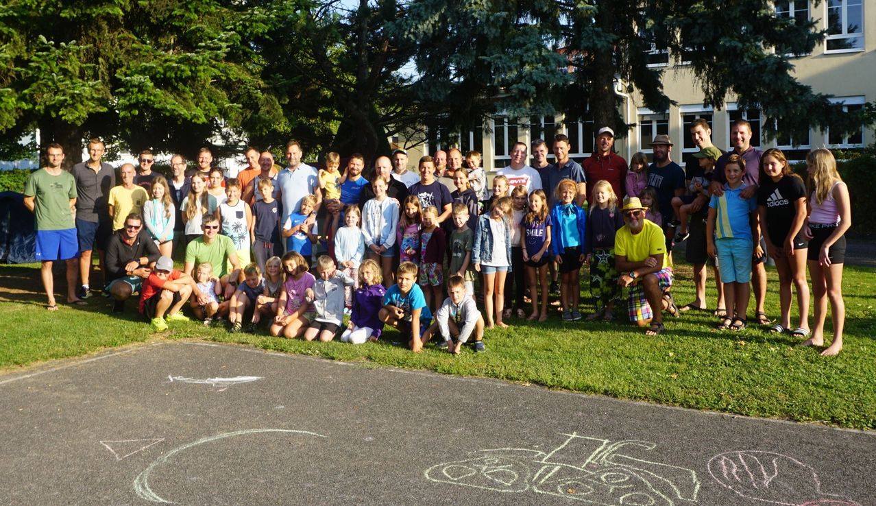 Eine Gruppe von Menschen, darunter Erwachsene und Kinder, posiert für ein Foto im Park. Bäume und ein Gebäude sind im Hintergrund zu sehen.