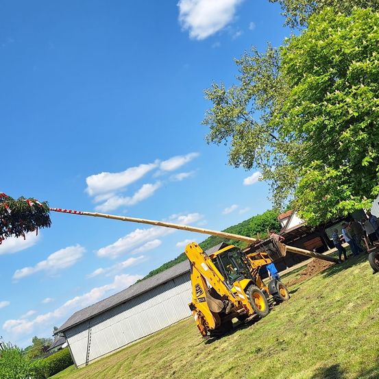 Ein gelber Bagger wird verwendet, um einen langen Holzbalken mit Bändern zu heben, unter einem blauen Himmel mit Wolken, in der Nähe eines Gebäudes und von Menschen.