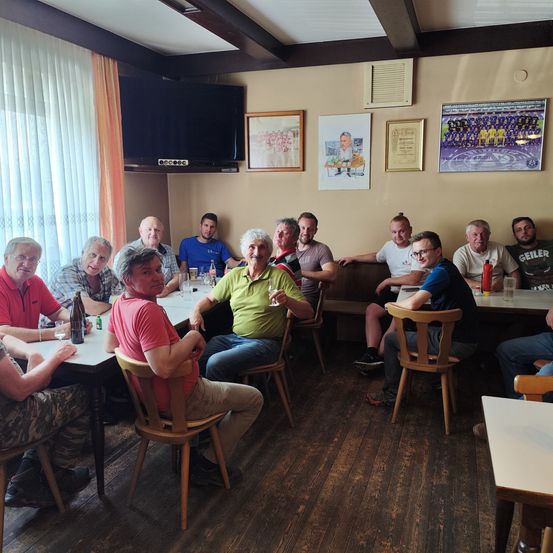 Eine Gruppe von Männern, die um Tische in einem Raum herumsitzen, einige mit Gläsern, andere mit Weinflaschen, und mehrere gerahmte Bilder an der Wand.