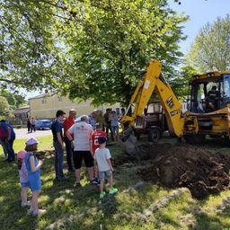 Eine Gruppe von Menschen versammelt sich unter einem Baum. Ein gelber Bagger gräbt ein Loch im Gras. Ein Mann sitzt im Bagger.