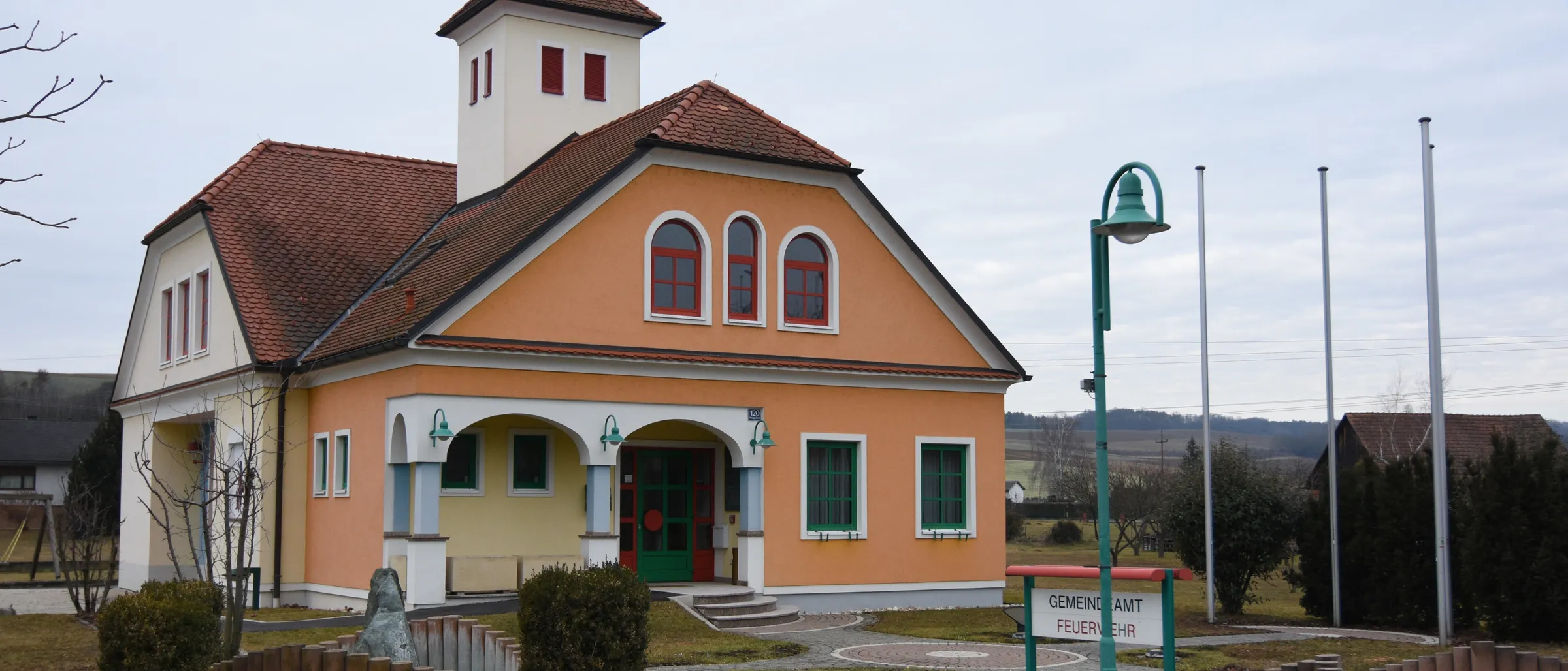Ein kleines Gebäude mit einem braunen Dach und orangefarbenen Wänden. Es hat einen Turm mit einer Glocke oben und mehrere Bogenfenster. Davor befindet sich eine Rasenfläche mit einem Brunnen und einem Schild, auf dem 'Gemeindeamt Feuerwehr' steht.