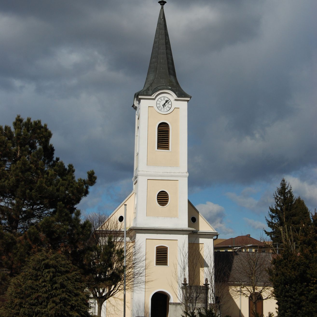 Eine beige Kirche mit einem hohen Turm und einer Uhr darauf steht zwischen Bäumen. Der Himmel darüber ist bewölkt.