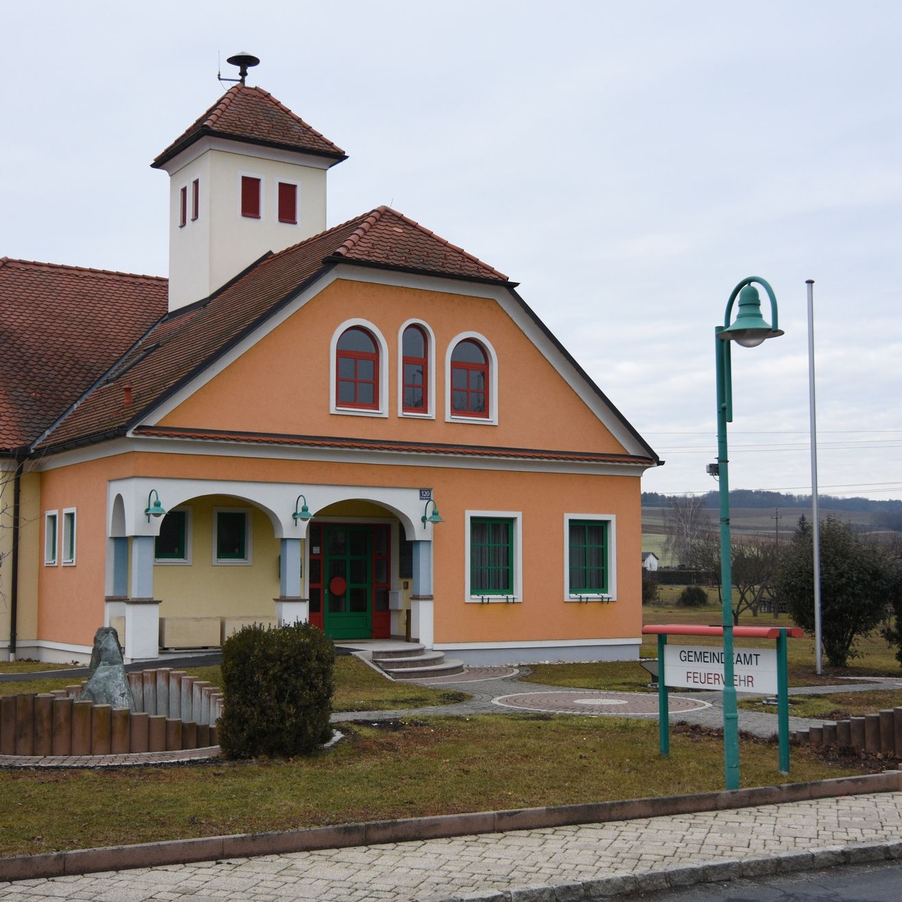 Ein kleines, zweistöckiges Gebäude mit braunem Dach und orangefarbenen Wänden, umgeben von Gras und einem kleinen Brunnen. Das Gebäude hat bogenförmige Fenster mit roten Rahmen und eine grüne Tür. Eine Straßenlampe ist in der Nähe.
