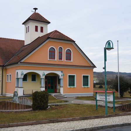 Ein kleines, zweistöckiges Gebäude mit braunem Dach und orangefarbenen Wänden, umgeben von Gras und einem kleinen Brunnen. Das Gebäude hat bogenförmige Fenster mit roten Rahmen und eine grüne Tür. Eine Straßenlampe ist in der Nähe.