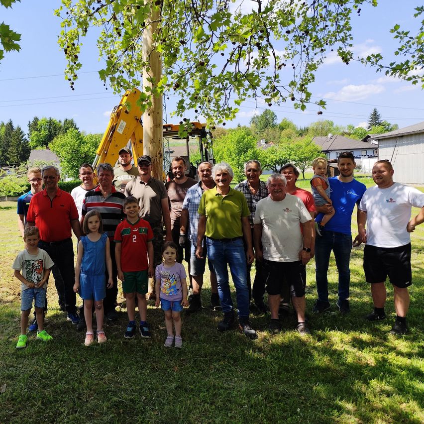 Eine Gruppe von Erwachsenen und Kindern, darunter ein Kind in einem roten Trikot, posiert für ein Foto in einem Garten. Im Hintergrund steht ein Baum und ein Bagger.