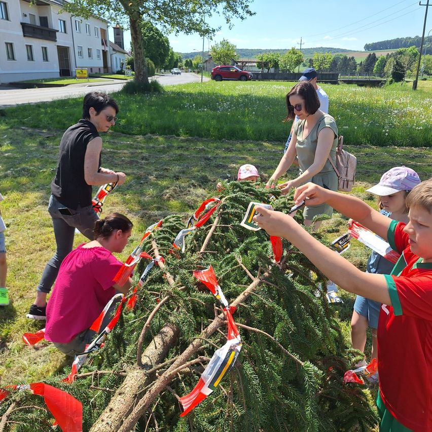 Eine Gruppe von Erwachsenen und Kindern schmückt gemeinsam einen großen immergrünen Baum mit roten und weißen dreieckigen Bändern.