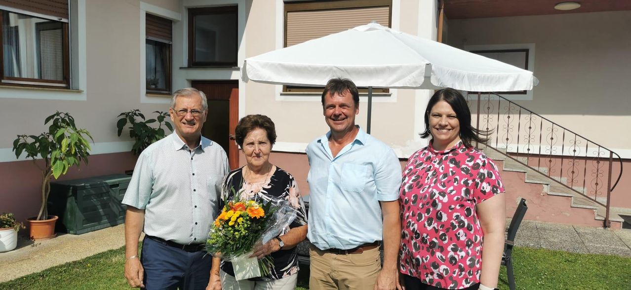 Vier Menschen stehen vor einem Haus mit einem weißen Sonnenschirm. Die Frau hält einen Blumenstrauß.