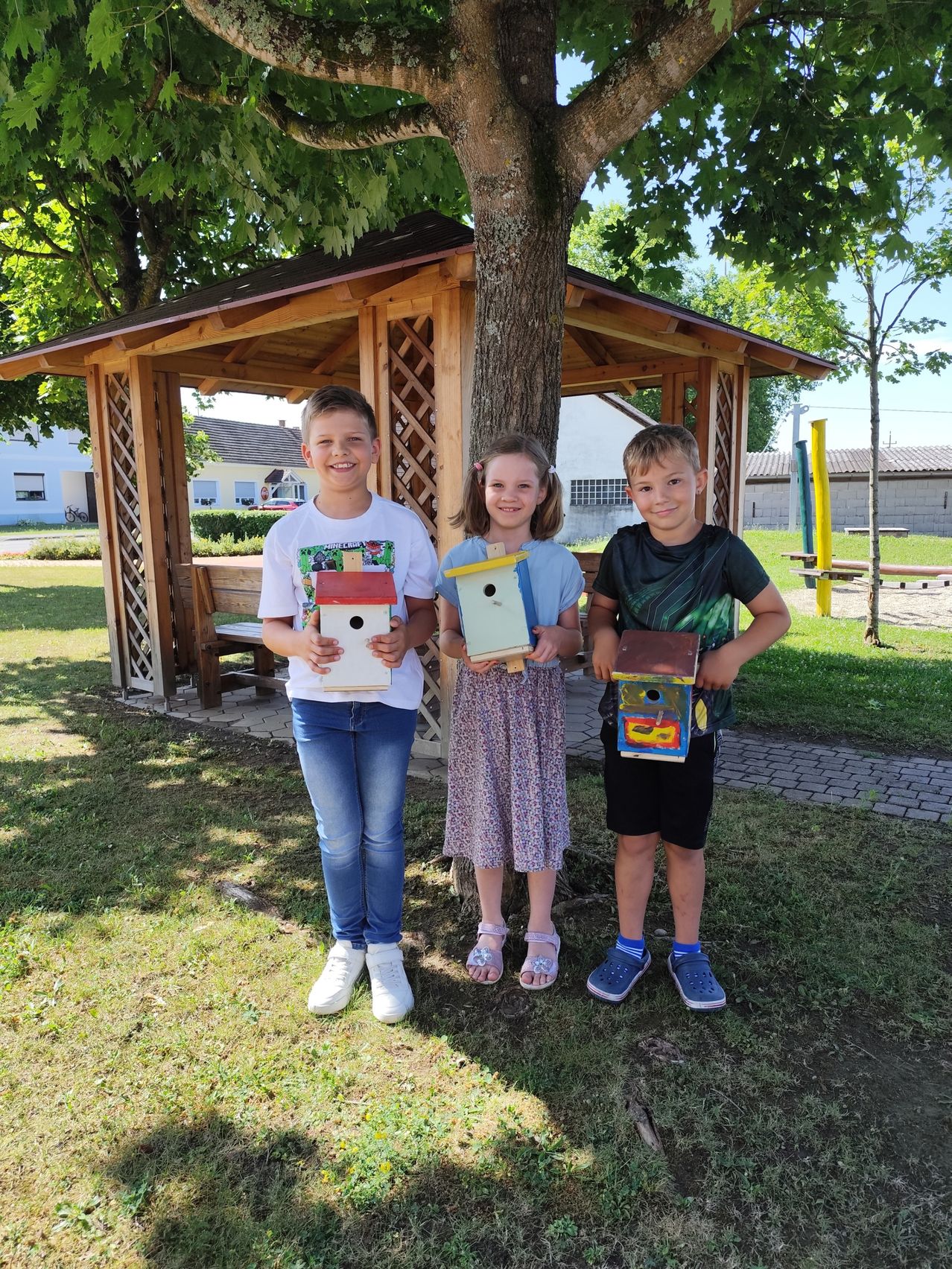 Drei Kinder stehen unter einem Baum, jedes hält ein bemaltes Vogelhaus. Sie lächeln für ein Foto in einem Park mit einem hölzernen Pavillon im Hintergrund.